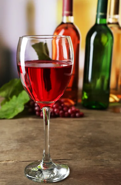 Copa de vino tinto con botellas de colores sobre mesa de madera sobre fondo desenfocado — Foto de Stock