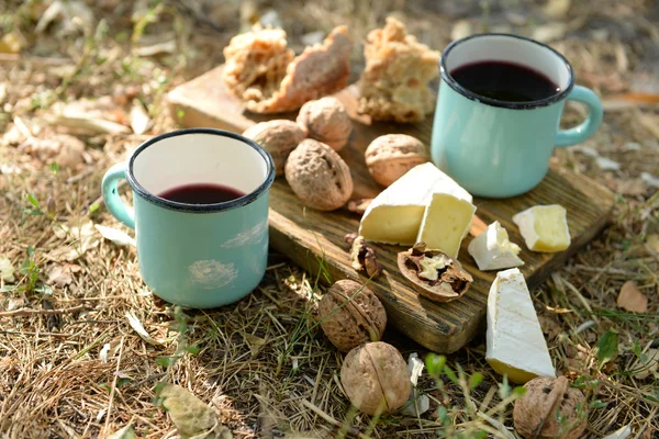 Tazze con vino rosso, deliziosi formaggi e noci su tavola di legno all'aperto - tema picnic — Foto Stock