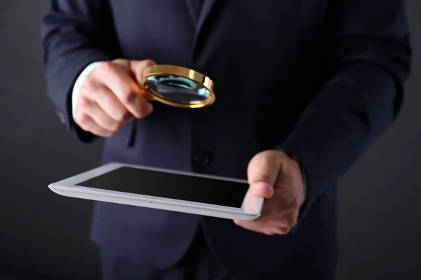 Geschäftsmann hält Lupe und digitales Tablet auf dunkelgrauem Hintergrund, sucht und begutachtet Konzept — Stockfoto