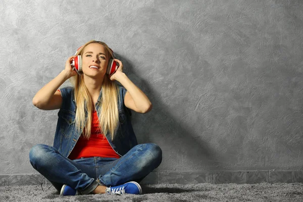 Young woman sitting on the floor and listening to music on a grey wall background — Stock Photo, Image