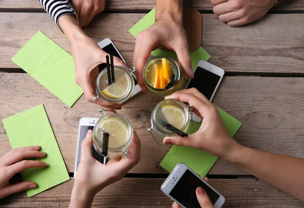 Cuatro manos con teléfonos inteligentes que sostienen cócteles, sobre fondo de mesa de madera — Foto de Stock