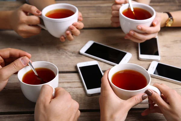 Quattro mani con smartphone che tengono tazze con tè, su sfondo tavolo di legno — Foto Stock