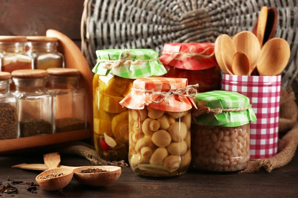 Gläser mit eingelegtem Gemüse, Bohnen, Gewürzen und Geschirr auf hölzernem Hintergrund — Stockfoto
