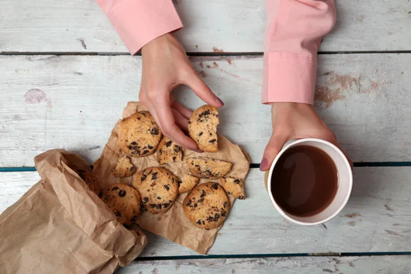 Kvinnliga händer som håller kopp kaffe och kakor på träbord nära håll — Stockfoto