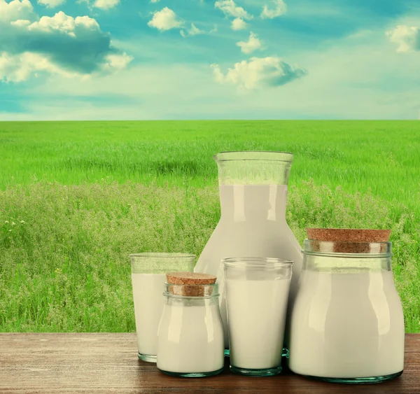 Jarra, frascos y vasos de leche sobre fondo de campo — Foto de Stock