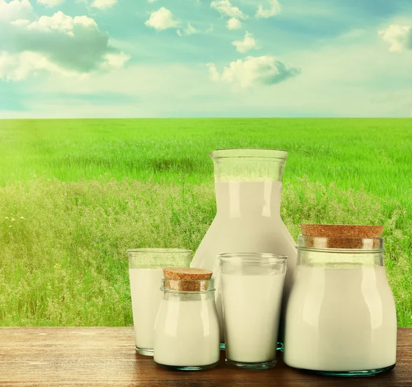 Jarra, frascos y vasos de leche sobre fondo de campo — Foto de Stock