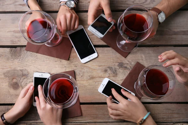 Four hands with smart phones holding glasses with red wine, on wooden table background — Stock Photo, Image