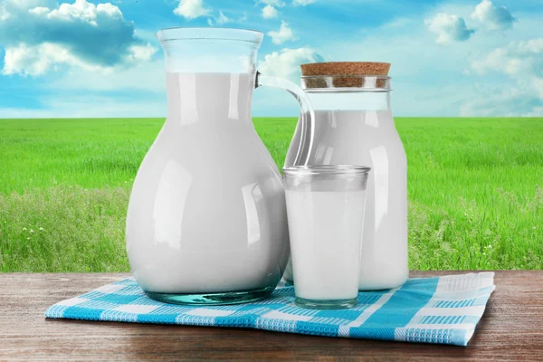 Pitcher, jar and glass of milk on wooden table, on nature background — Stock Photo, Image