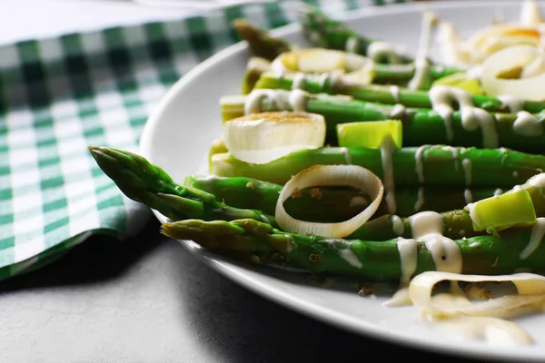 Fresh asparagus dish with sliced onion on white plate with checkered cotton serviette on grey table, close up — Stock Photo, Image