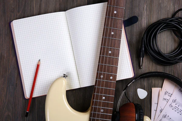 Electric guitar with headphones, musical notes and note book on wooden background