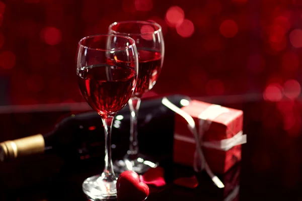 Setting of glasses with wine, bottle and a gift in the box, on red blurred background — Stock Photo, Image
