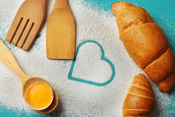 Heart of flour, croissant and  wooden kitchen utensils on color wooden background — Stock Photo, Image