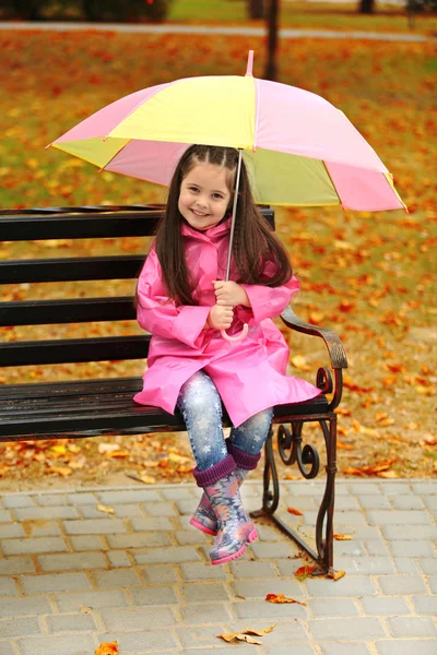 Vacker liten flicka med paraply sitter på bänken i park — Stockfoto