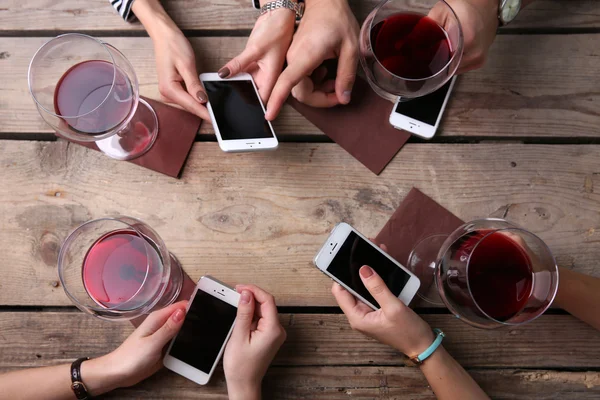 Four hands with smart phones holding glasses with red wine, on wooden table background — Stock Photo, Image