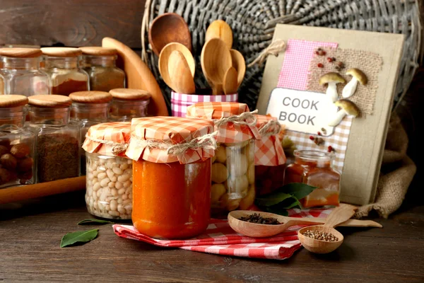 Frascos com legumes em conserva e feijão, especiarias, livro de receitas e utensílios de cozinha sobre fundo de madeira — Fotografia de Stock