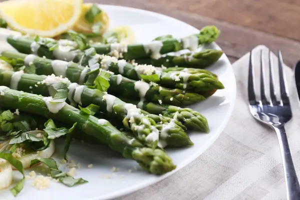 Appetizing asparagus in sour cream sauce with onion, cheese and lemon on white plate against wood background, close up — Stok Foto