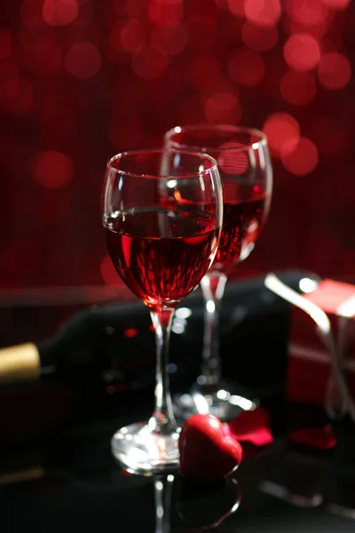 Ajuste de copas con vino, botella y un regalo en la caja, sobre fondo rojo borroso —  Fotos de Stock