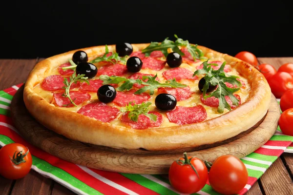 Tasty pizza with salami and olives decorated with tomatoes on wooden table against black background — Stock Photo, Image