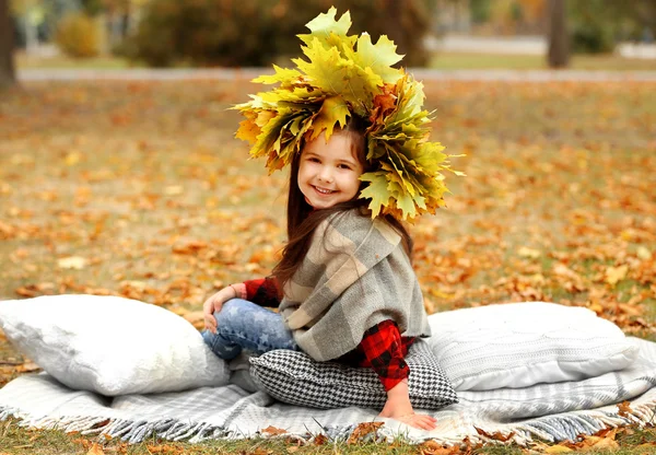 Gelukkig jong meisje in gele herfst krans zittend op plaid in park — Stockfoto