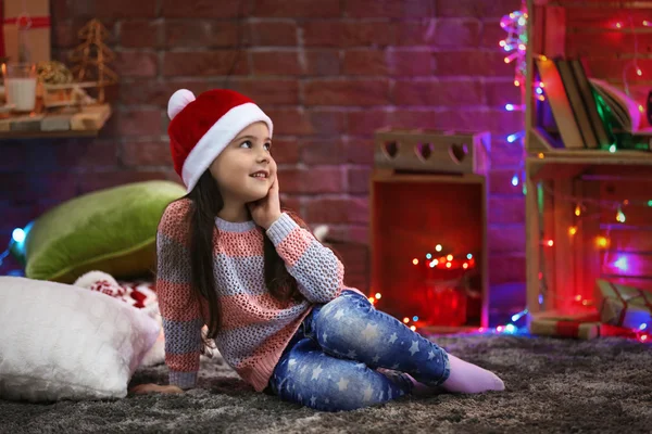 Pretty little girl in red hat sitting in Christmas decorated room — Stock Photo, Image