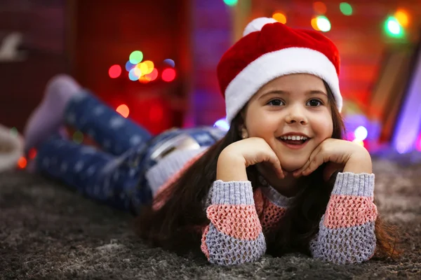 Pretty little girl in red hat laying in Christmas decorated room — Stock Photo, Image