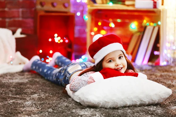 Hübsches kleines Mädchen mit rotem Hut liegt auf dem Kissen im weihnachtlich dekorierten Zimmer — Stockfoto