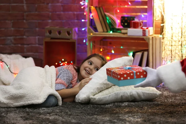 Hübsches kleines Mädchen liegt auf dem Kissen unter weichem Karo und wartet auf das Weihnachtsgeschenk im weihnachtlich dekorierten Zimmer — Stockfoto