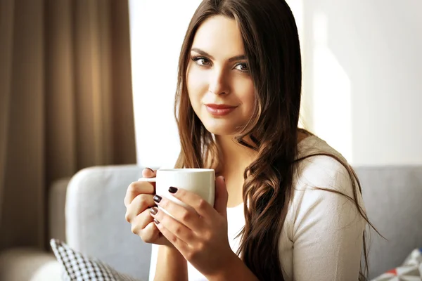 ผู้หญิงสวยผ่อนคลายบนโซฟาในห้องพร้อมถ้วยกาแฟ — ภาพถ่ายสต็อก