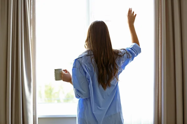 Femme avec une tasse de café regardant par la fenêtre dans la chambre — Photo