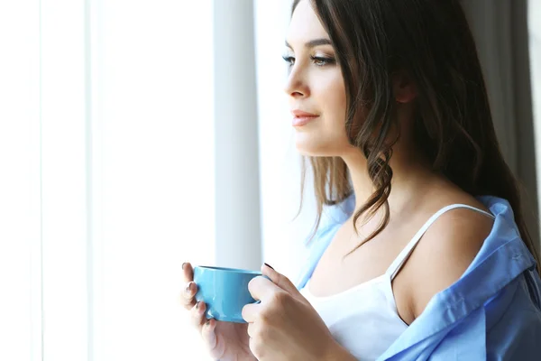 Donna con tazza di caffè in piedi vicino alla finestra nella stanza — Foto Stock