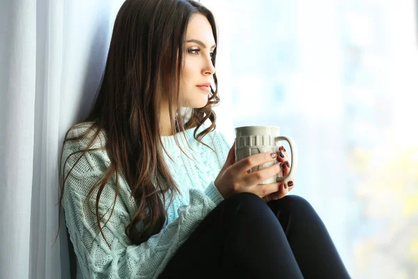 Mujer bonita sentada en el tablero de la ventana con una taza de café — Foto de Stock