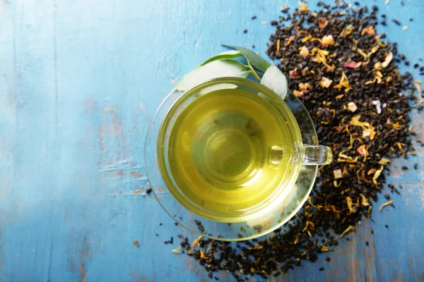 Glas kopje thee met groene bladeren en verspreide thee rond op blauwe houten achtergrond — Stockfoto