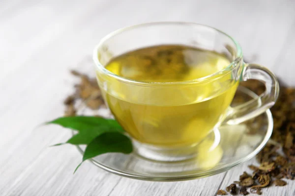 Стеклянная чашка чая с зелеными листьями и разбросанный чай вокруг на сером деревянном фоне, закрыть — стоковое фото