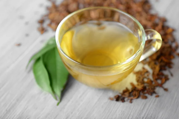 Glass cup of tea with green leaves and scattered tea around on grey wooden background, close up — Stock Photo, Image