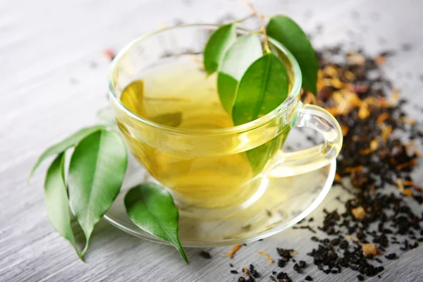 Стеклянная чашка чая с зелеными листьями и разбросанным чаем на сером деревянном фоне — стоковое фото