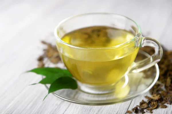 Copa de té de vidrio con hojas verdes y té disperso sobre fondo de madera gris, de cerca — Foto de Stock