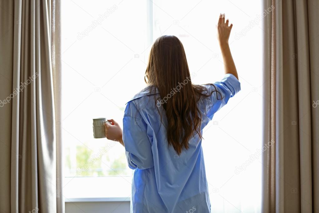 Woman with cup of coffee looking through the window in the room