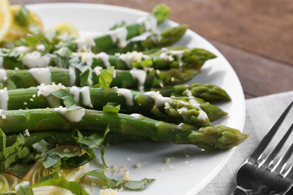 Smakelijk asperges in zure roomsaus met ui, kaas en citroenschil op wit bord tegen houten achtergrond, close-up — Stockfoto