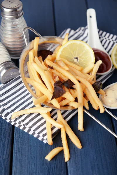 French fried potatoes in metal colander with sauce on wooden background — Stock Photo, Image