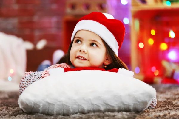 Pretty little girl in red hat laying on the pillow in Christmas decorated room — Stock Photo, Image