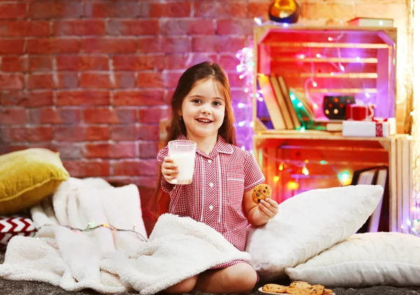 Красивая маленькая девочка со стаканом молока и вкусным печеньем в украшенной рождественской комнате — стоковое фото