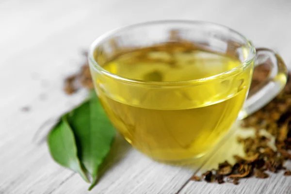 Copa de té de vidrio con hojas verdes y té disperso sobre fondo de madera gris, de cerca — Foto de Stock