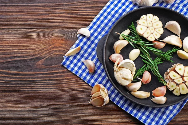 Fresh garlic with rosemary in black pan near blue checkered cotton napkin on wooden background — Stock Photo, Image