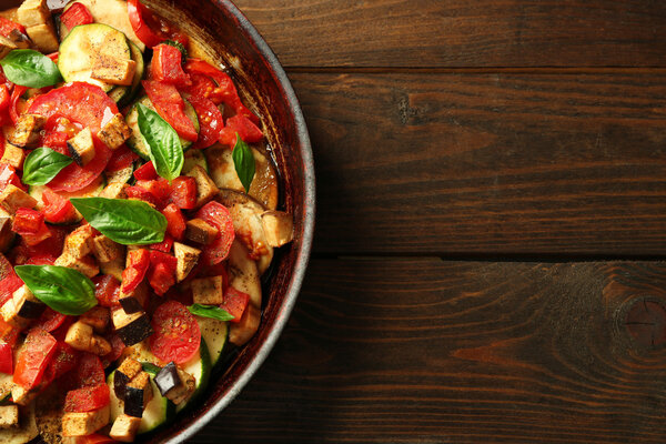 Tasty vegetarian ratatouille made of eggplants, squash, tomatoes and onions in black cast iron pan, on wooden table background