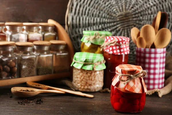 Jars with pickled vegetables and beans, spices and kitchenware on wooden background — Stock Photo, Image