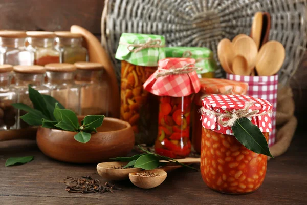 Jars with pickled vegetables, beans, spices and kitchenware on wooden background — Stock Photo, Image