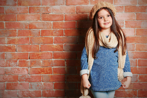 Portrait of little fashion kid girl in knitted cap and scarf on bricks wall background