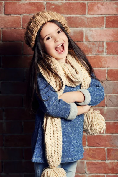 Retrato de pequena menina de moda em boné de malha e cachecol no fundo da parede de tijolos — Fotografia de Stock