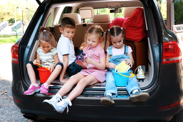 Tres hermosas chicas y un niño se sientan en el maletero de un coche y se ríen —  Fotos de Stock