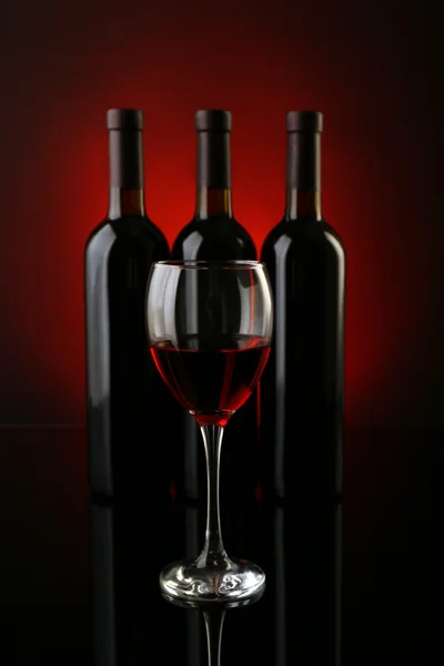 Wine glass against bottles in a row on dark red background, close up — Stock Photo, Image